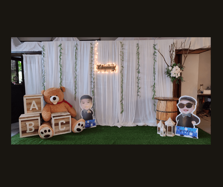 White draped baby celebration backdrop with hanging photos, greenery, toys, and letter blocks