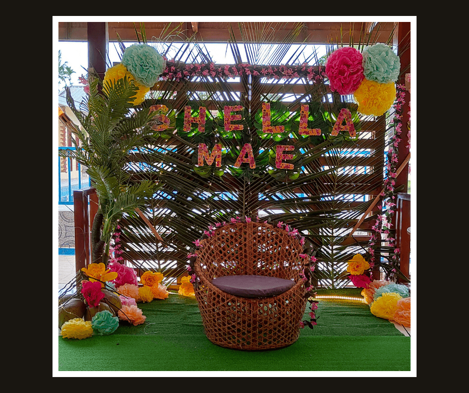Dark timber slat wall with colorful tropical flowers, palm leaves, paper fans, and woven seating