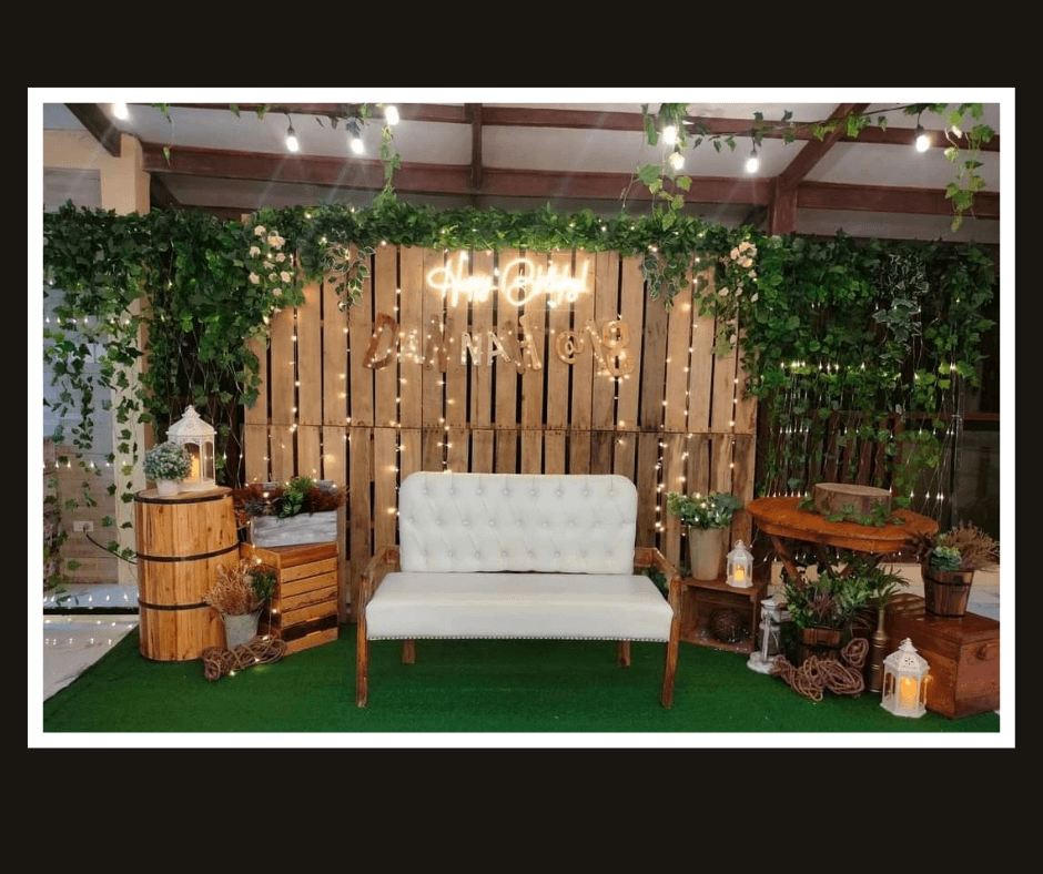 Rustic vertical plank backdrop framed with dense greenery, barrels, and a white loveseat