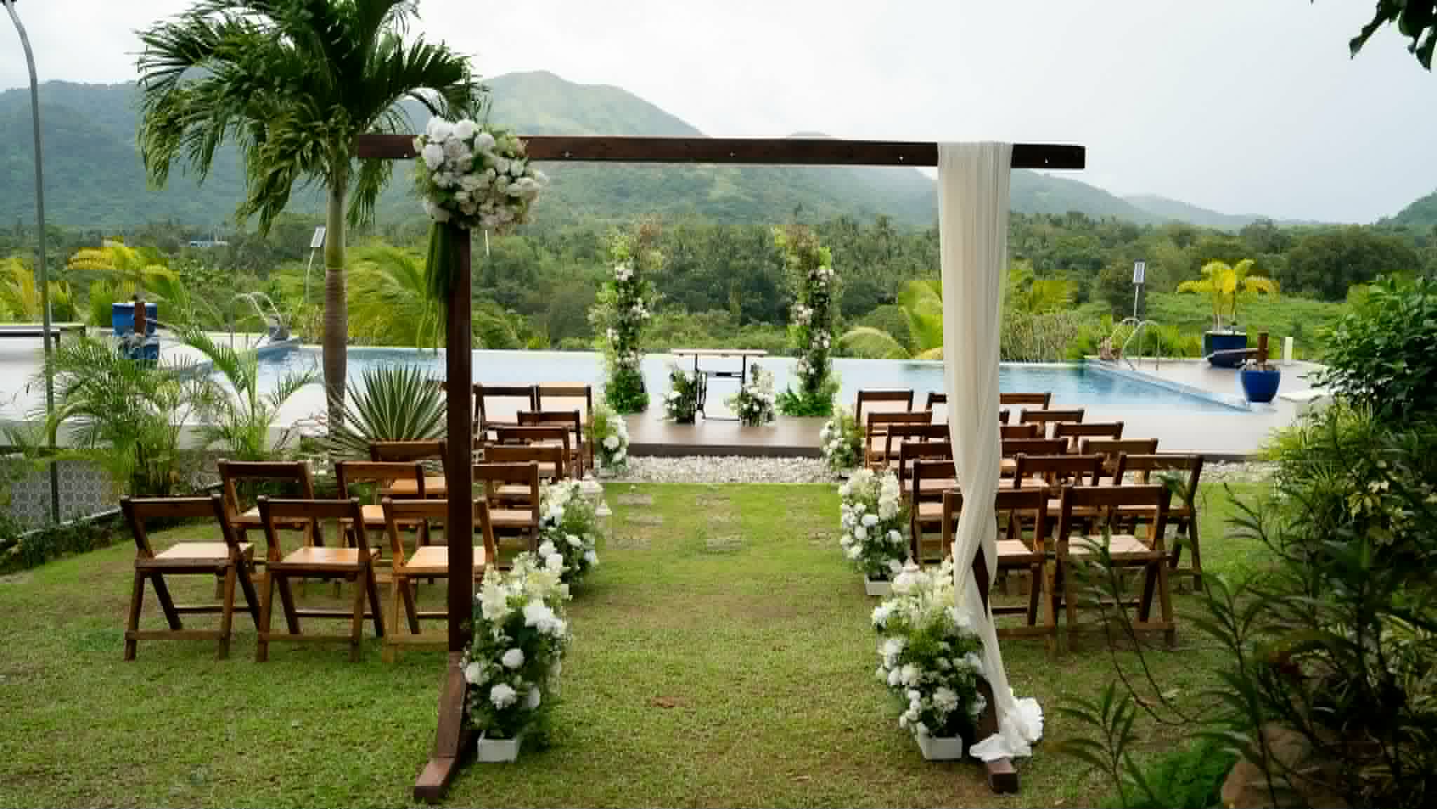 Natural wood ceremony arch framing a garden aisle beside a pool