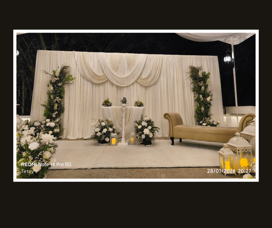 Indoor white draped backdrop styling with greenery panels, flowers, a sofa, and a round table