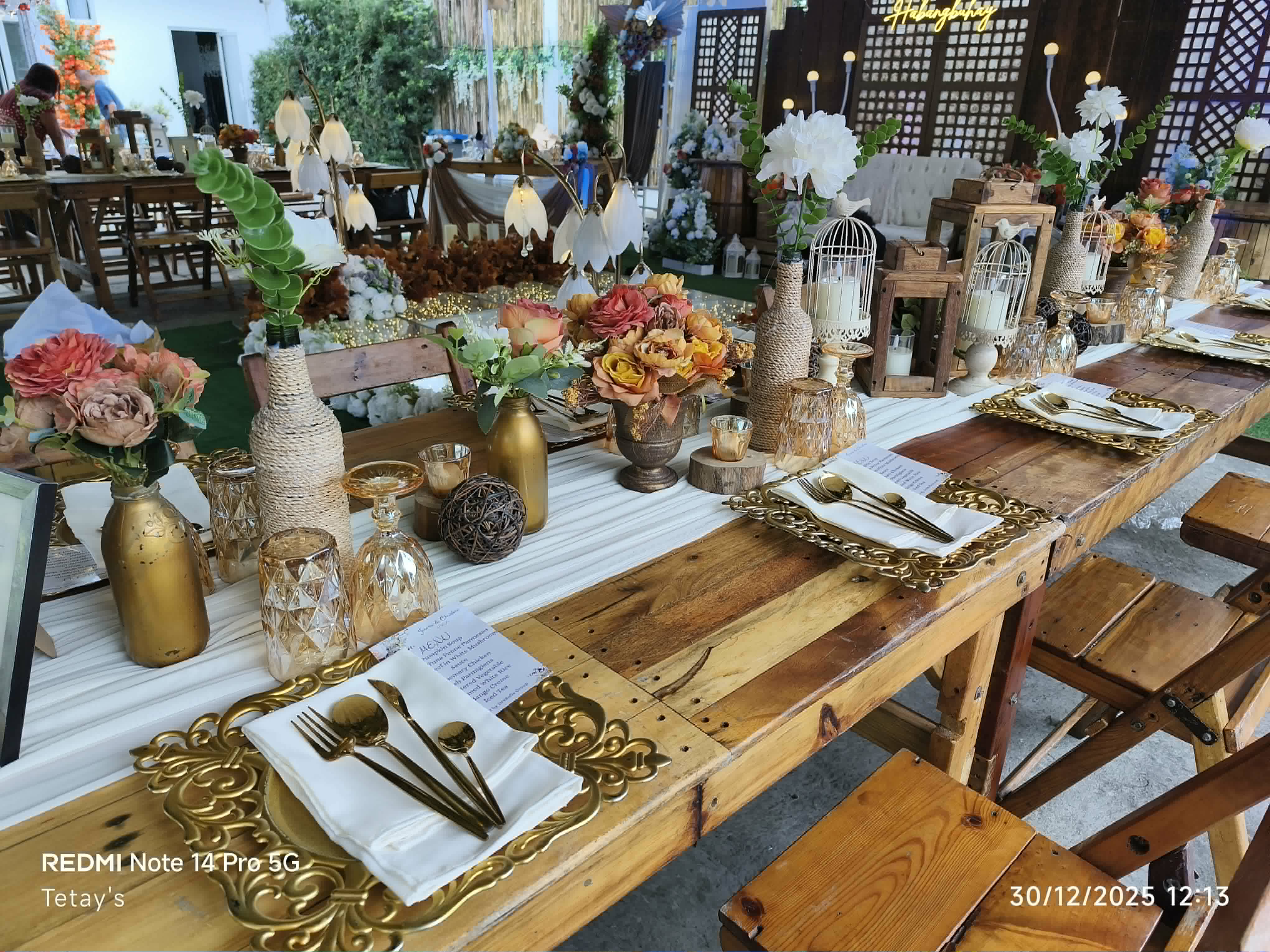 Rustic wooden reception table styled with gold bottles, flowers, lanterns, and place settings
