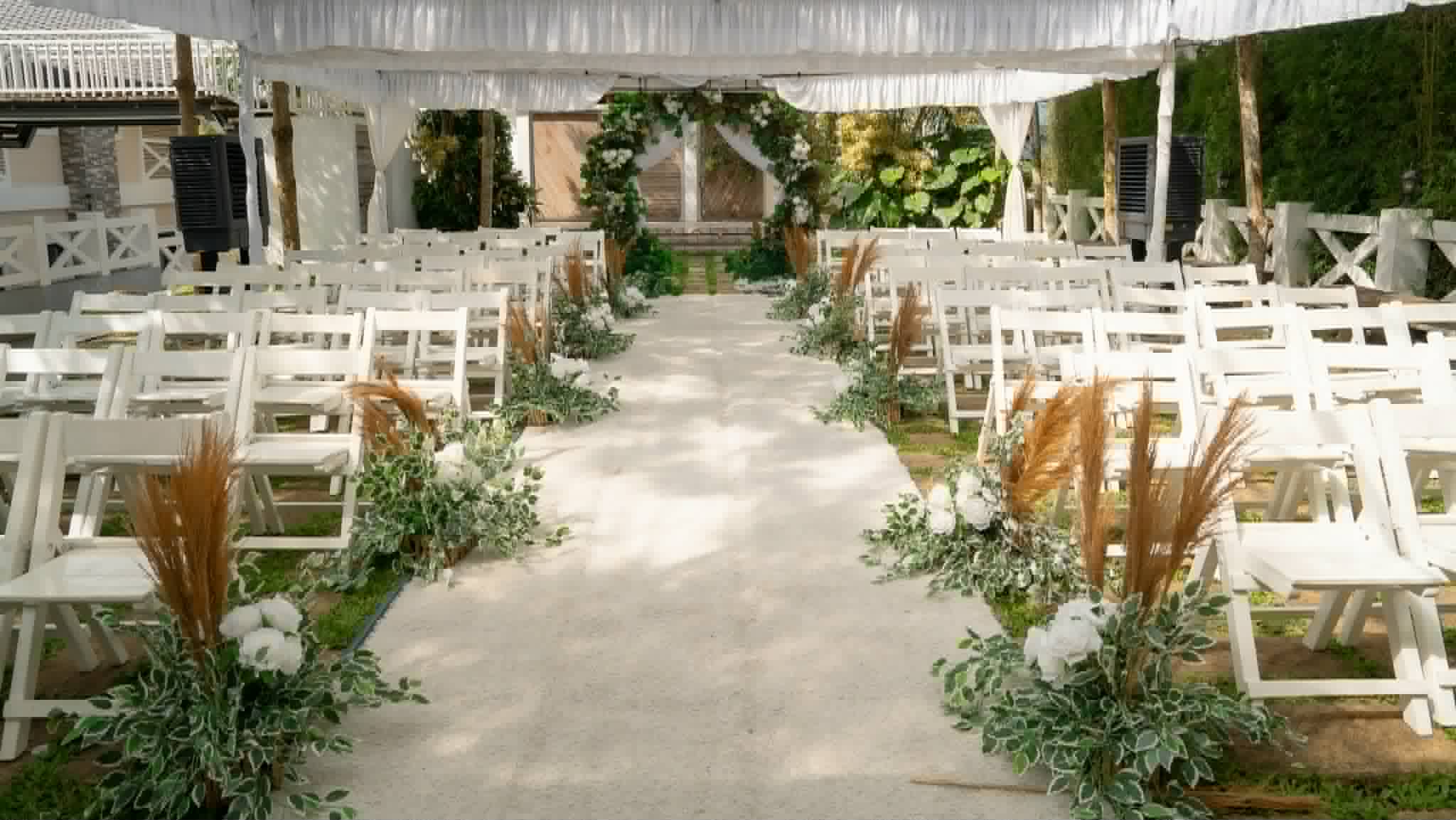 White wooden chairs arranged along a flower-lined aisle beneath a wedding tent